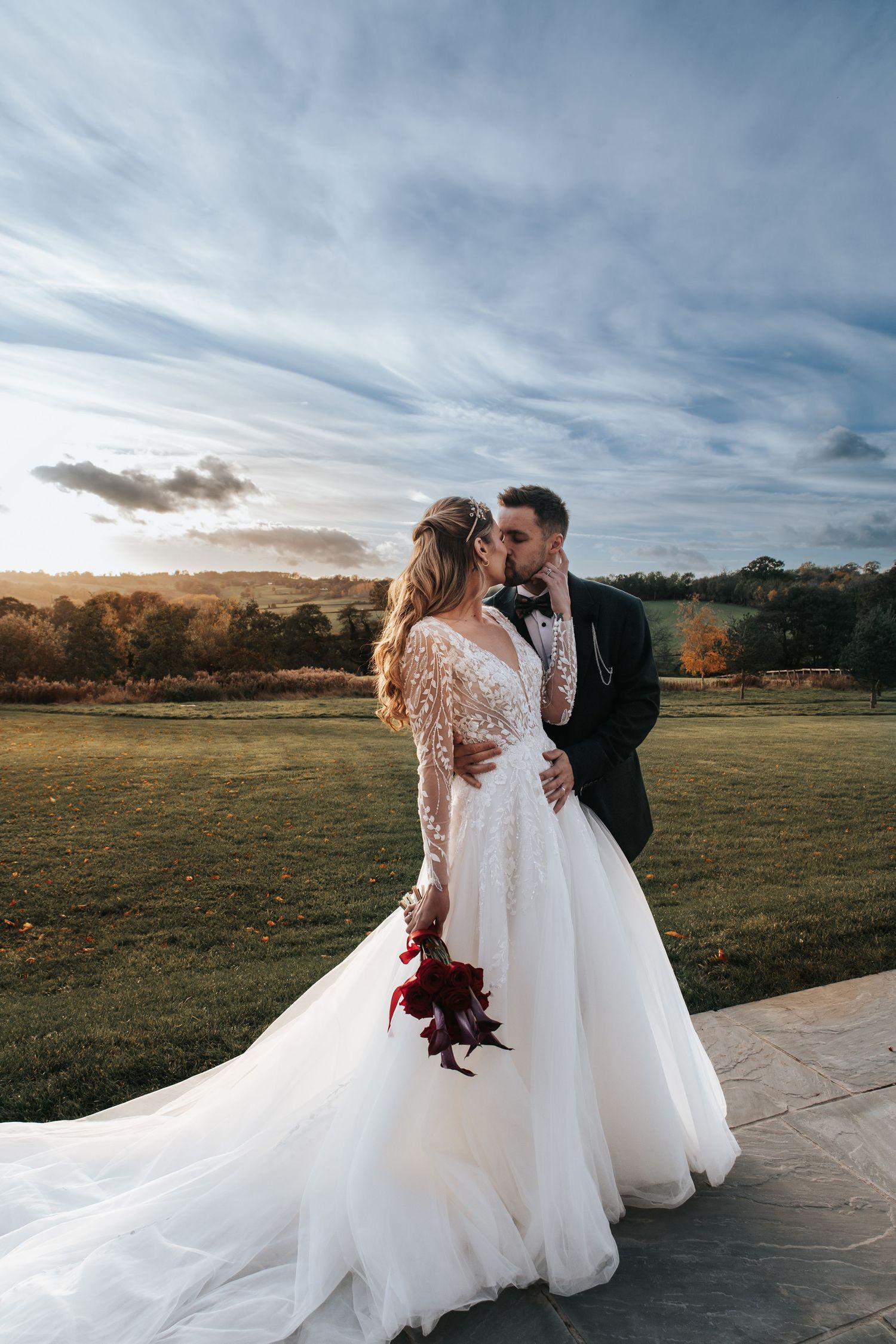 Romantic wedding couple embracing at sunset in a rustic outdoor field with dramatic cloudy sky.