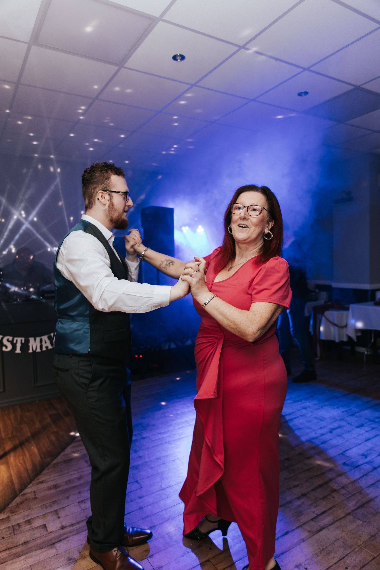 Two people dancing together on a wooden dance floor with blue lighting and a festive atmosphere in the background.