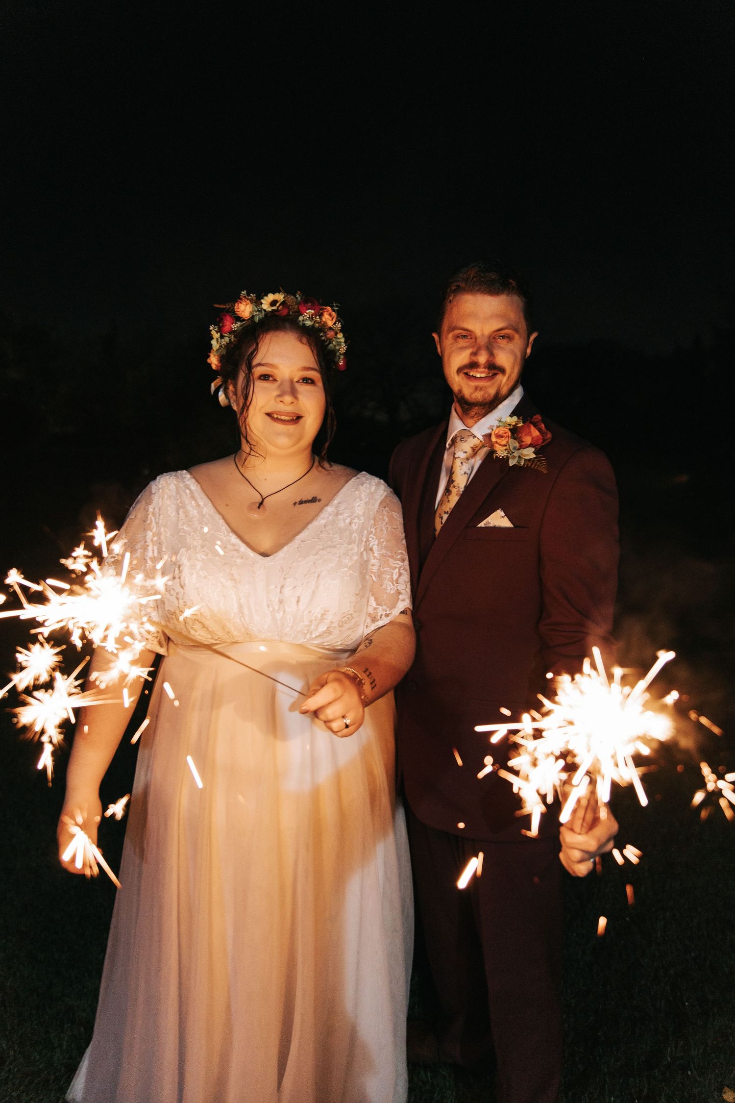 A couple holds sparklers while celebrating their wedding outdoors at night in a romantic setting.