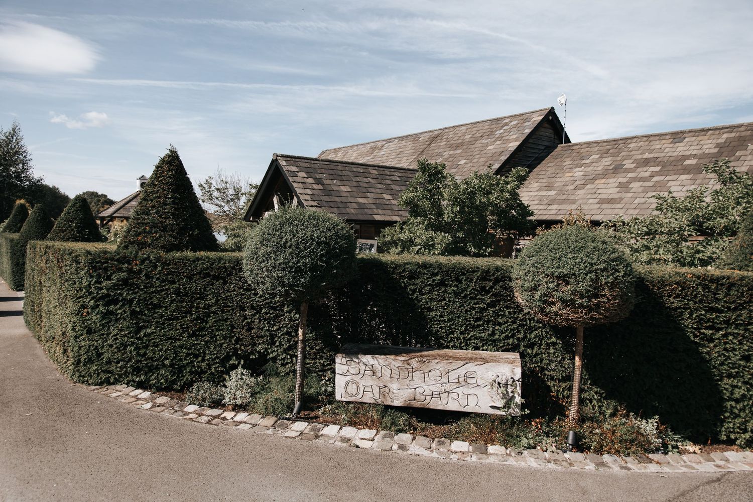 Rustic barn venue with manicured topiary hedges and a welcome sign, surrounded by lush greenery under a blue sky.