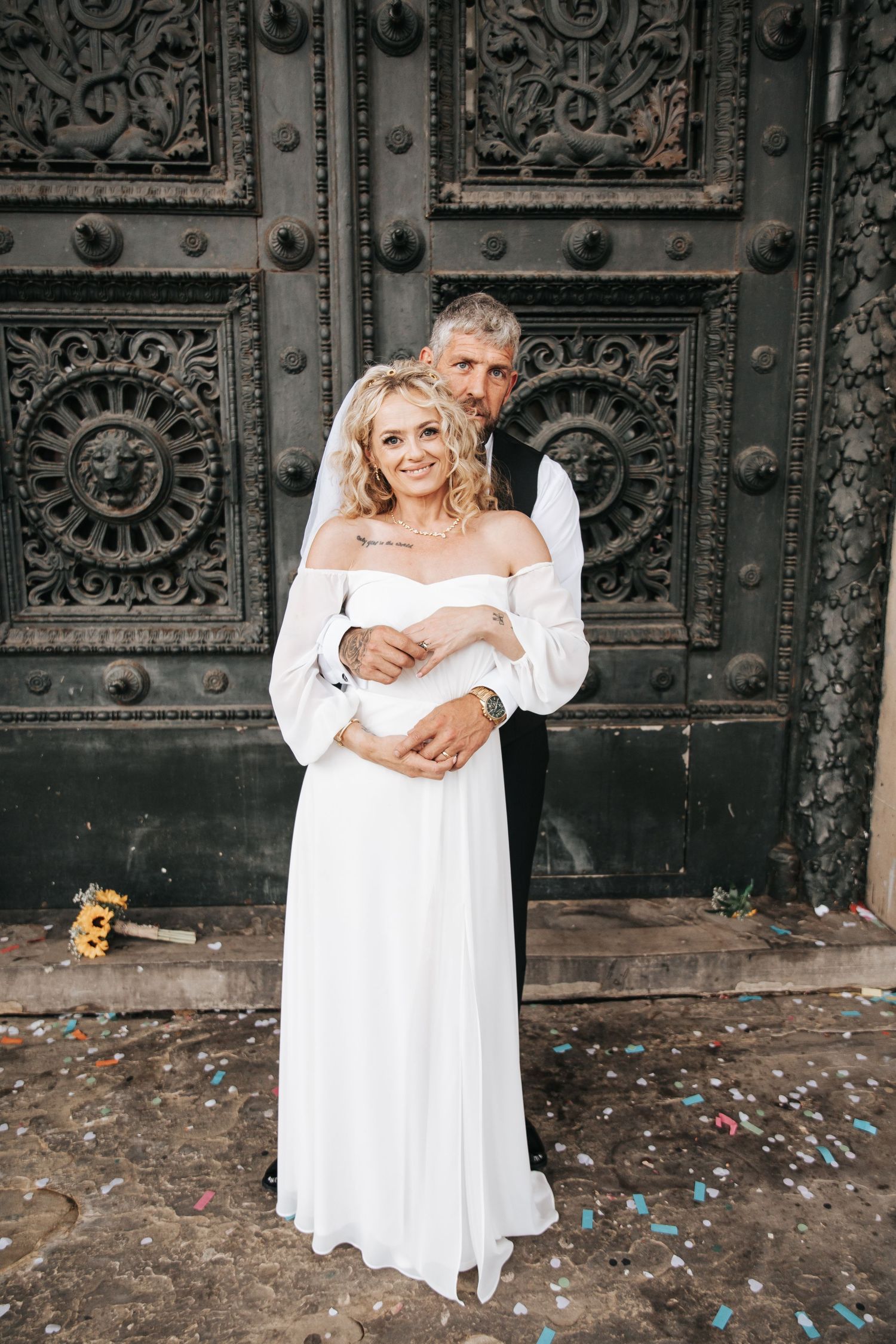 A smiling bride in white gown and veil embraced by groom before ornate dark metal doors with confetti on ground.
