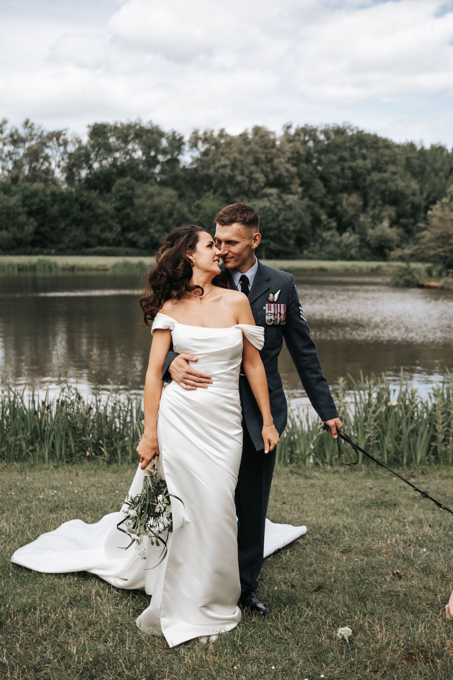 Military groom in uniform with medals kisses bride in white off-shoulder gown by a serene lake surrounded by lush trees.