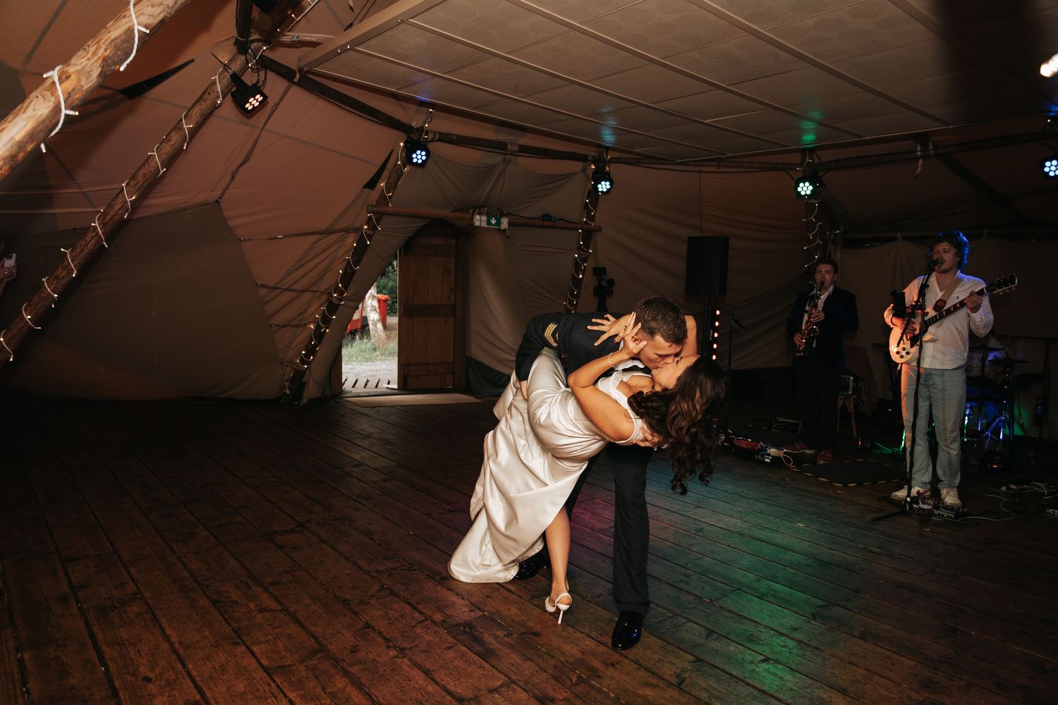 Bride and groom share romantic first dance dip in a marquee tent with live band performing in background.