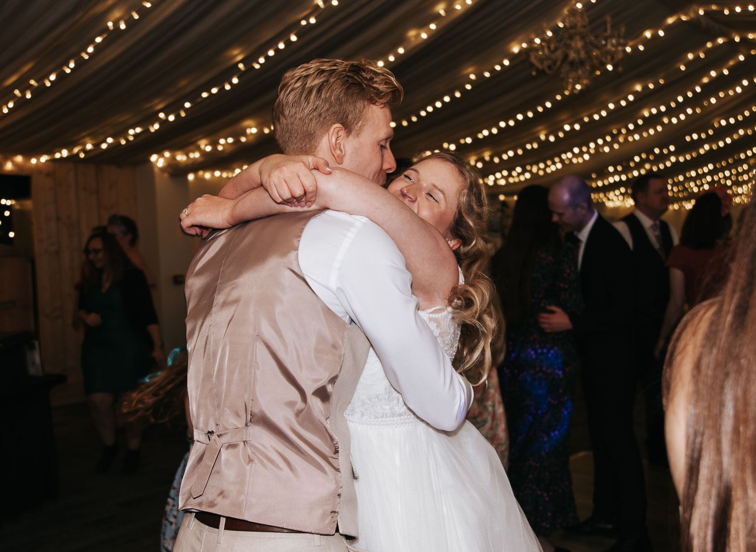 Bride and groom share first dance embrace under warm string lights at romantic wedding reception.