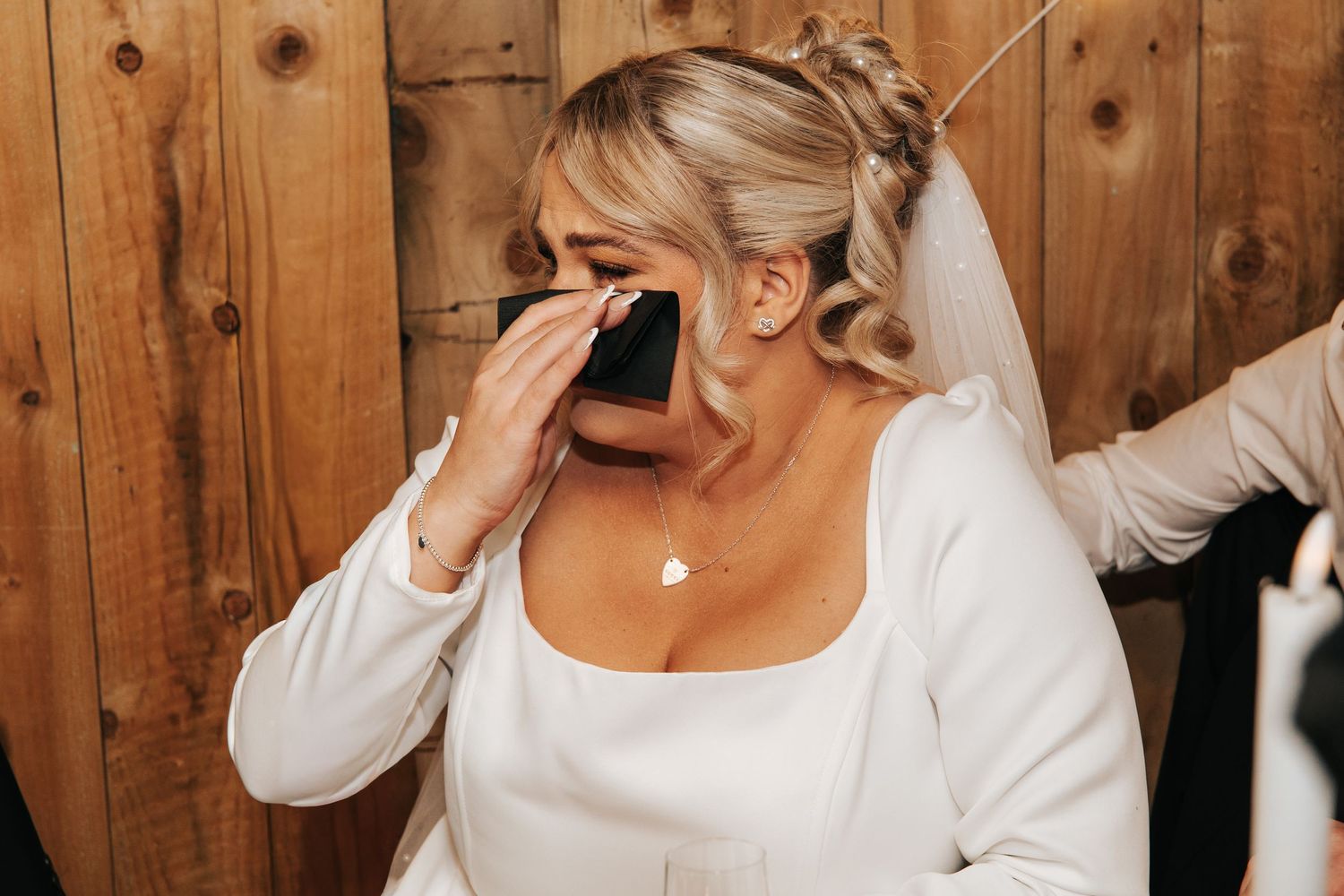 Emotional bride in white dress wipes tears during touching wedding moment against wooden backdrop.