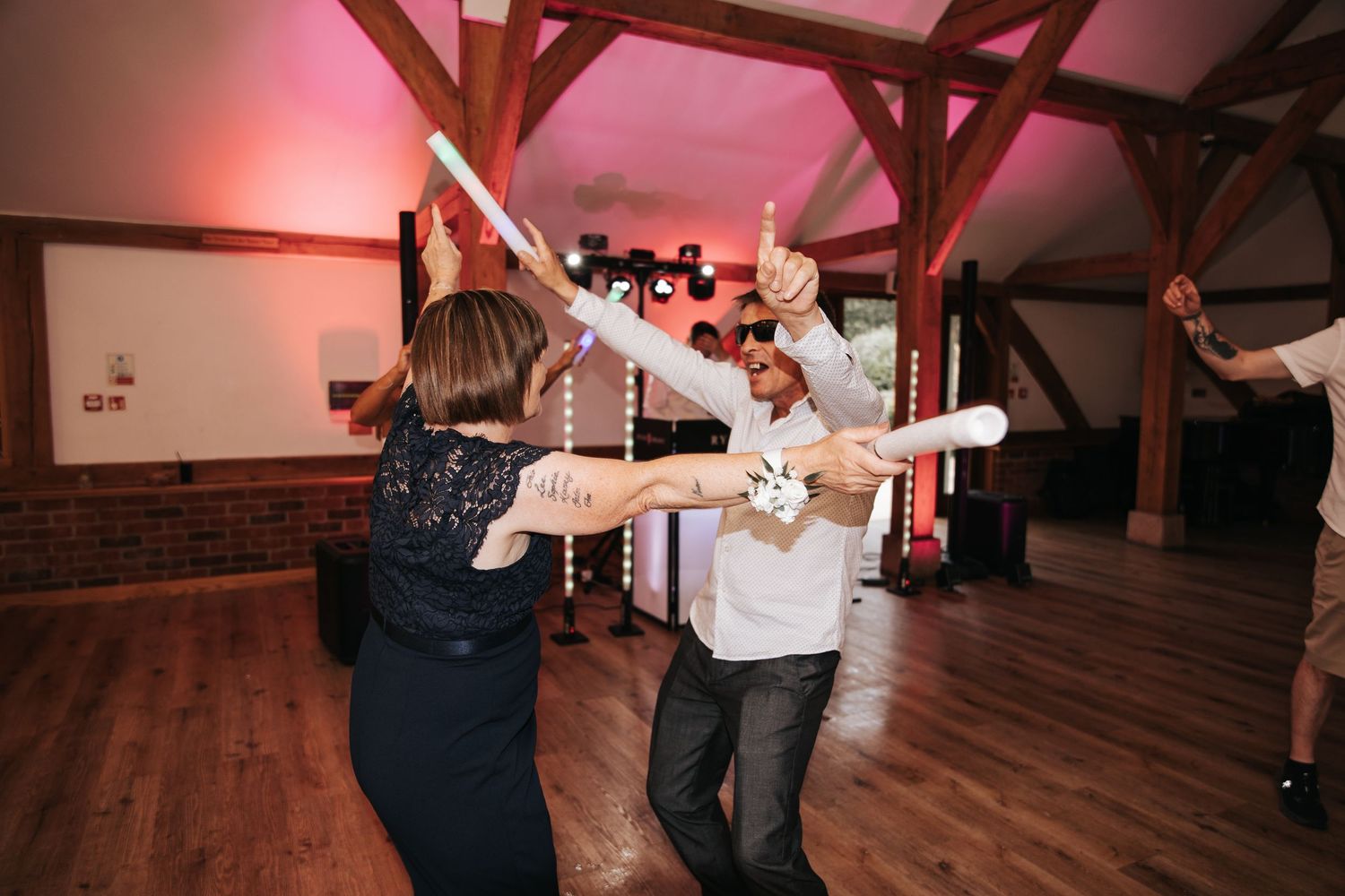 Wedding guests dancing with glow sticks at a rustic barn reception venue with warm pink lighting.
