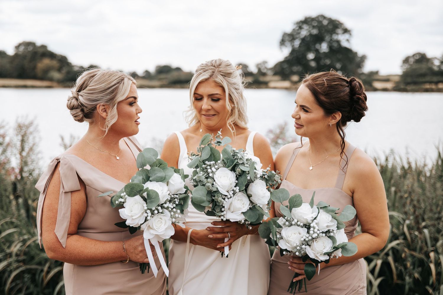 Bride in white gown with two bridesmaids in champagne dresses holding white and eucalyptus bouquets by a lake.