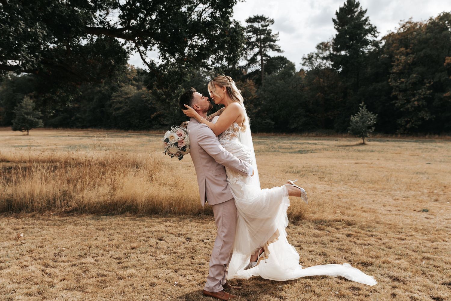 Groom lifts bride in white gown during outdoor wedding photoshoot in golden field surrounded by trees.