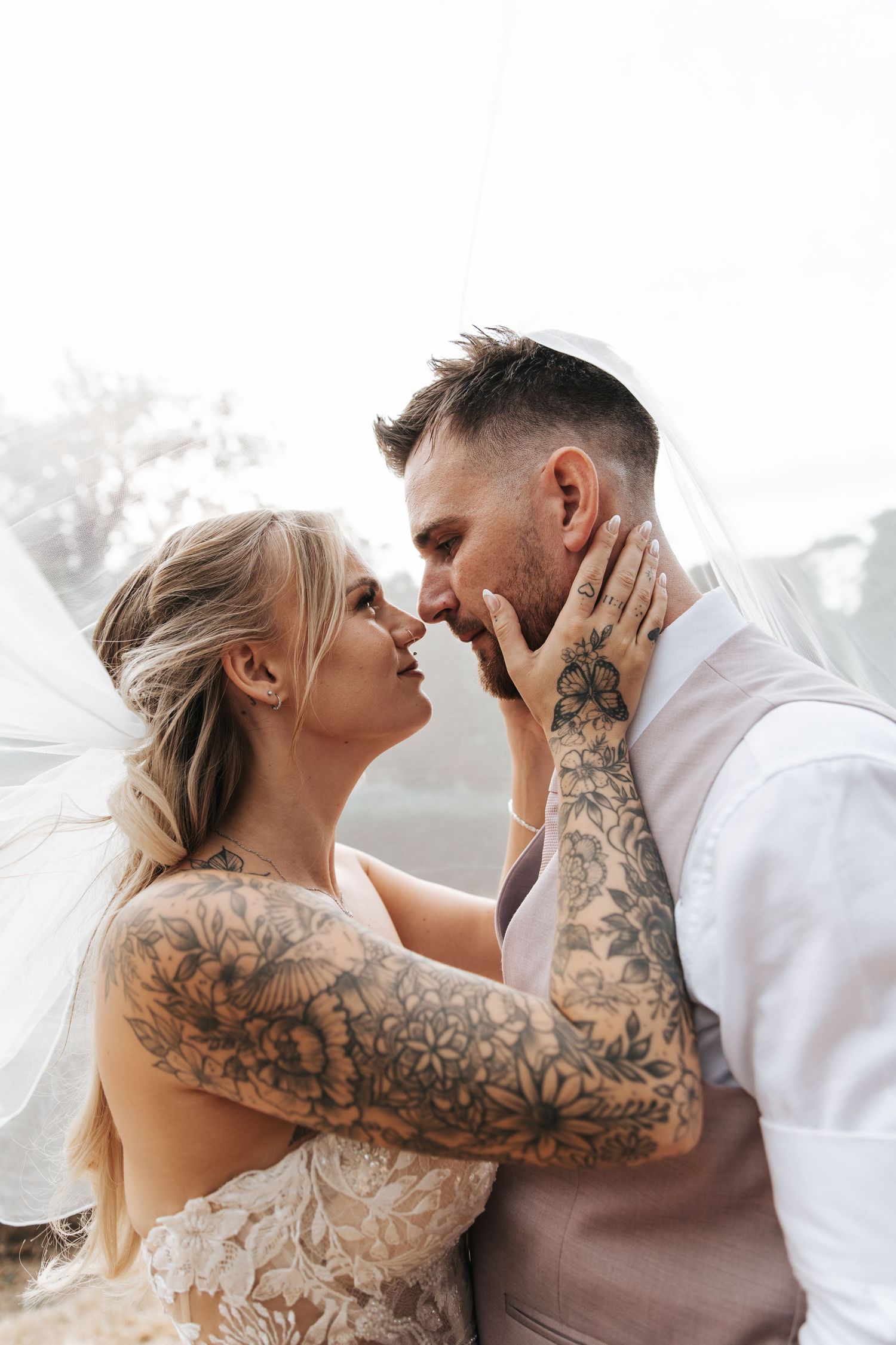 Tattooed bride and groom share a romantic almost-kiss under a veil on their wedding day.