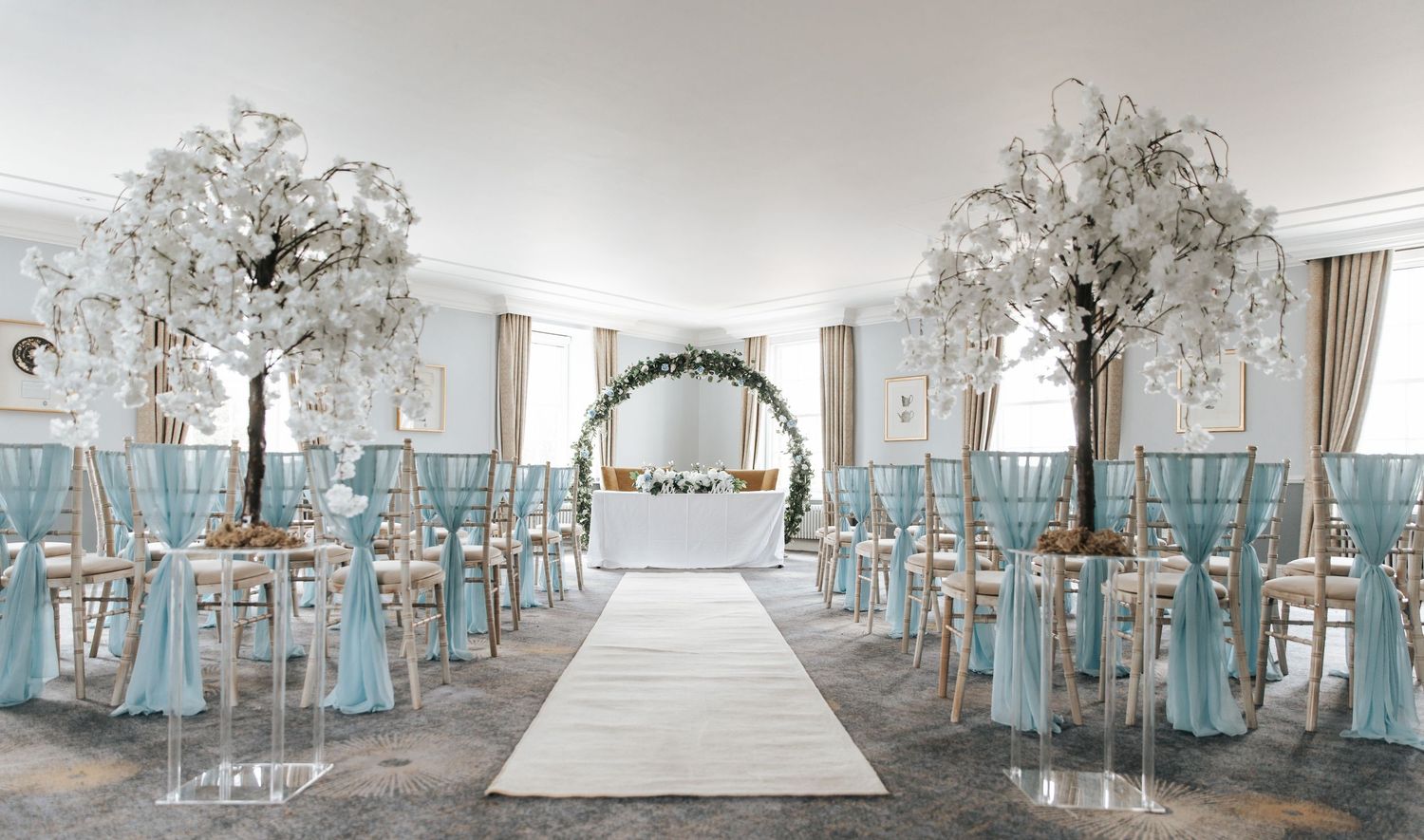 Elegant wedding ceremony setup with blue satin chair sashes, white floral trees, and a floral arch altar.