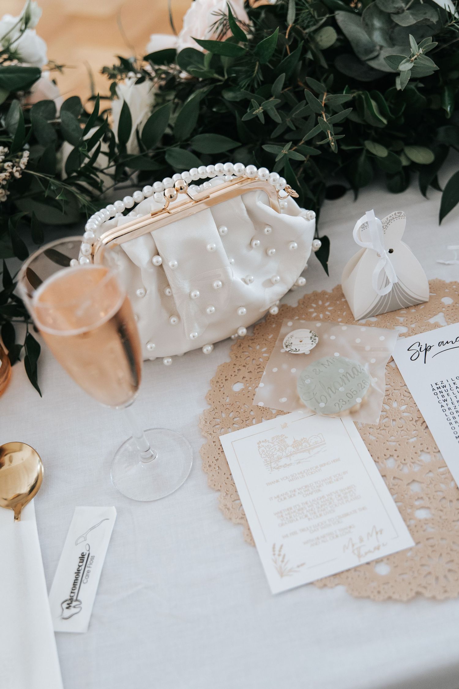 Elegant wedding table with pearl clutch bag, champagne flute, menu card, and lush greenery centerpiece.