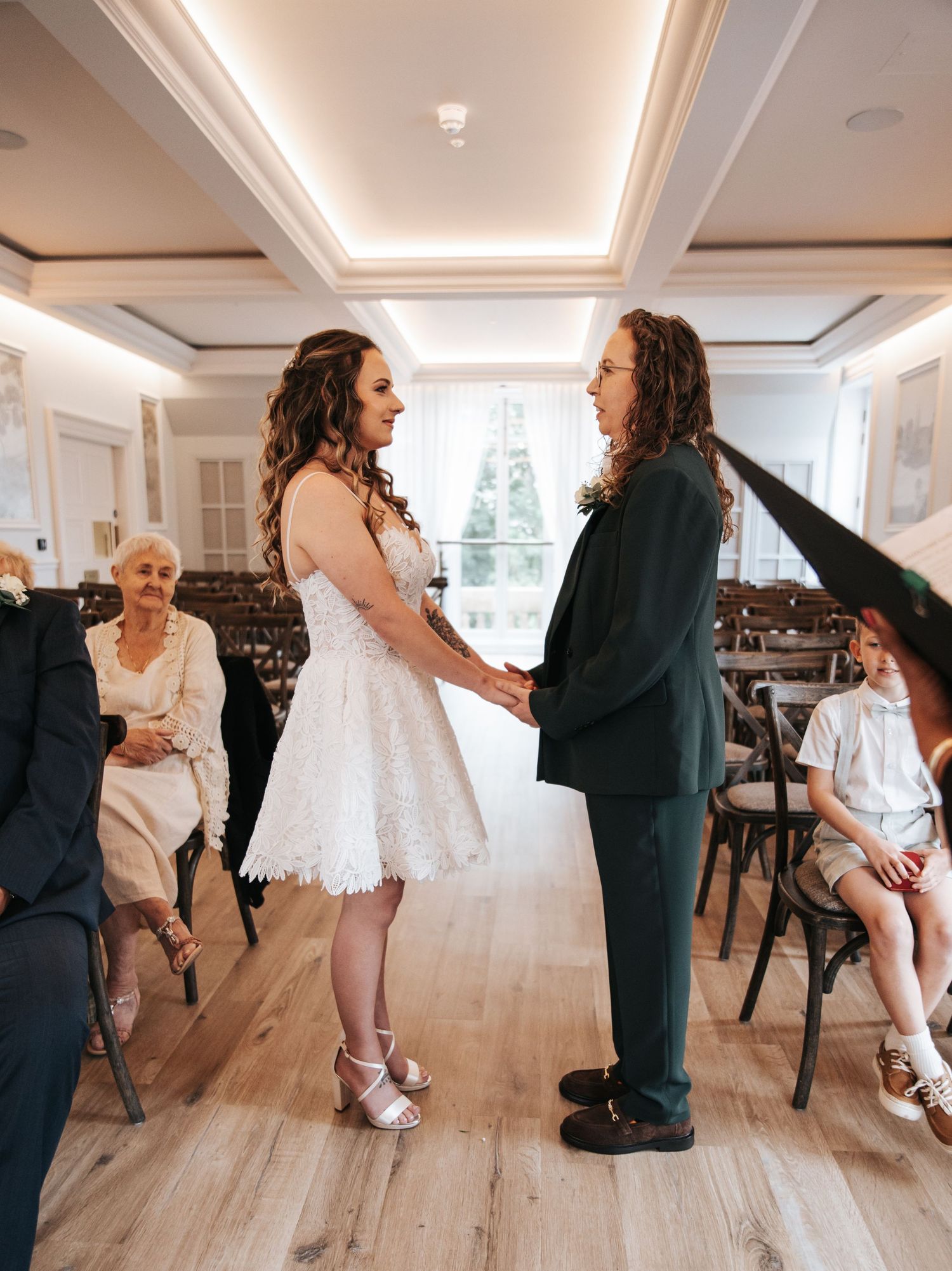 Couple holding hands during indoor wedding ceremony, bride in short lace dress, groom in dark suit, guests seated nearby.