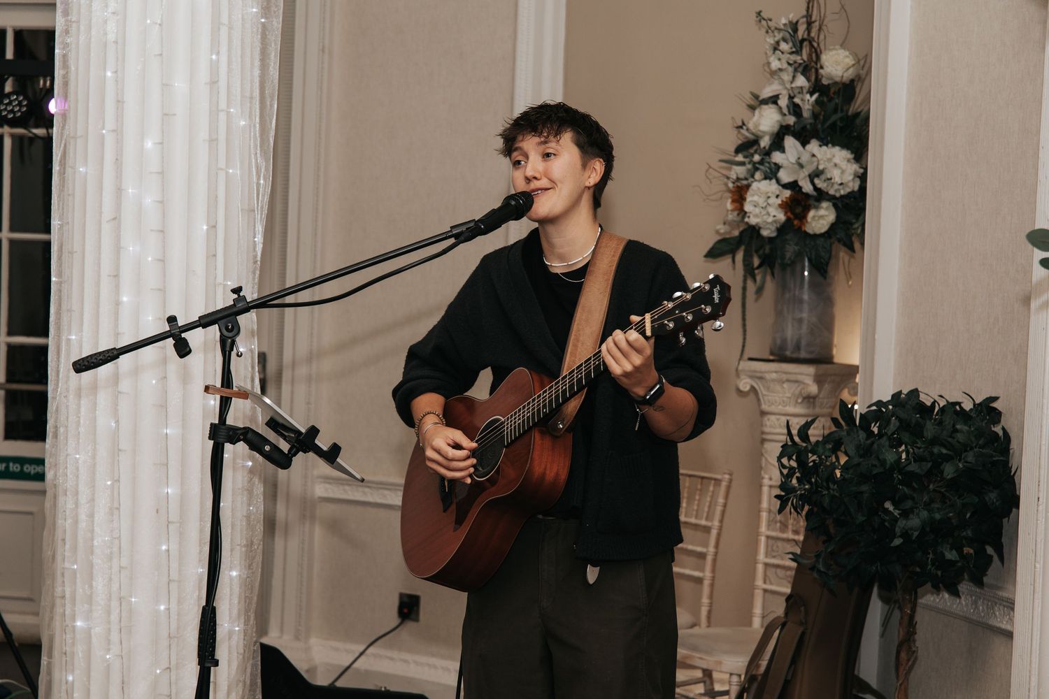 Musicians perform with acoustic guitars at an elegant indoor venue decorated with white lights and floral arrangements.