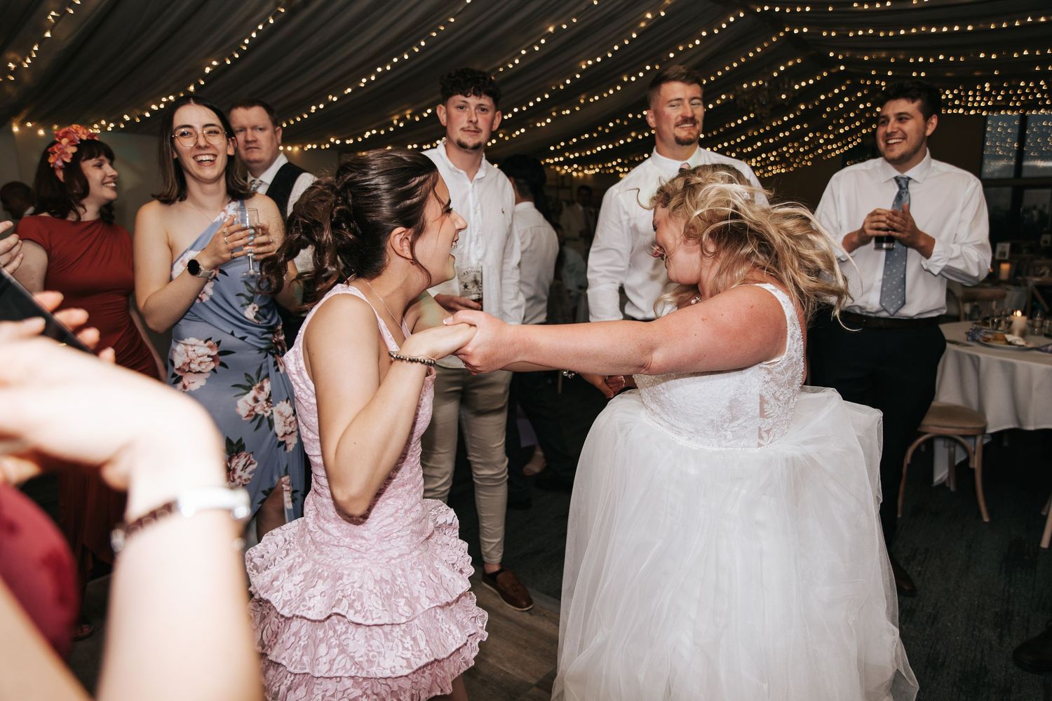 Bride and guest dancing joyfully at wedding reception under fairy lights.