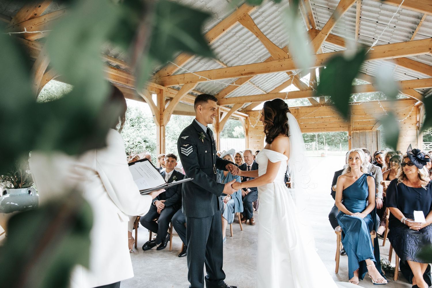 Military groom and bride exchange vows at an elegant outdoor wedding ceremony under a wooden pavilion with seated guests.