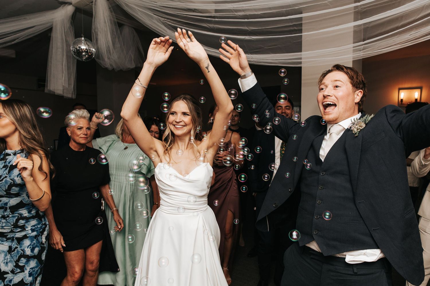 Joyful bride in white gown and groom in black suit celebrating at wedding reception with bubbles and guests.