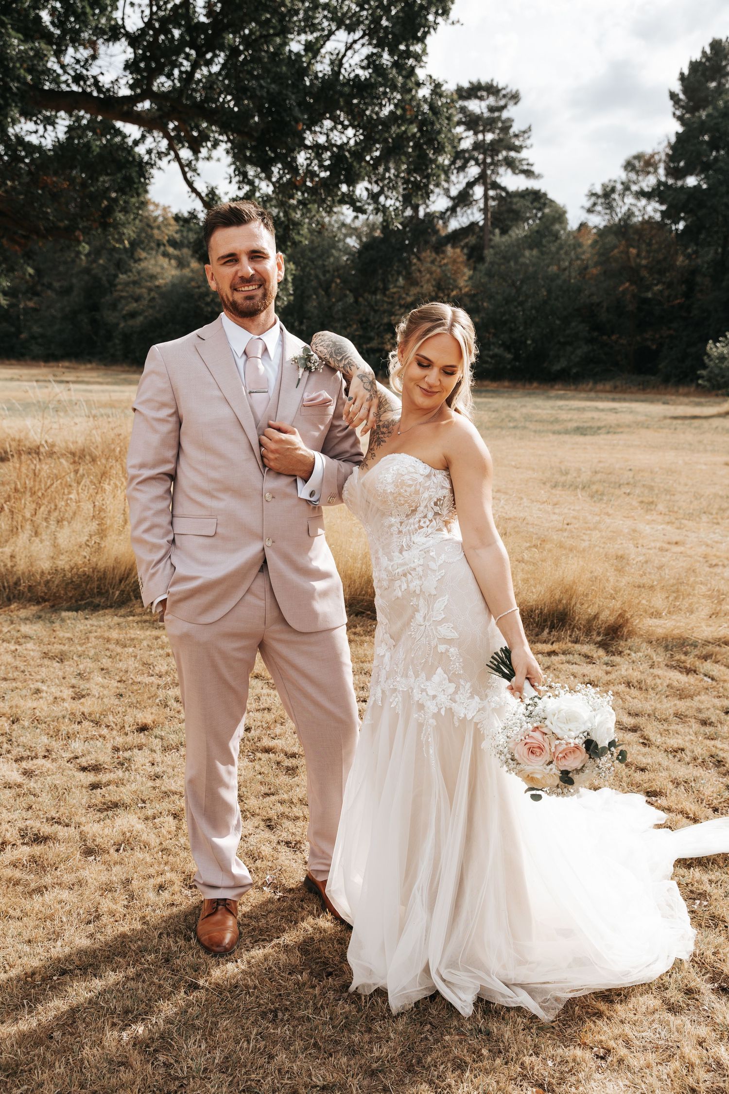 A smiling groom in a blush suit stands with his bride in a lace wedding gown holding a bouquet in a golden field.