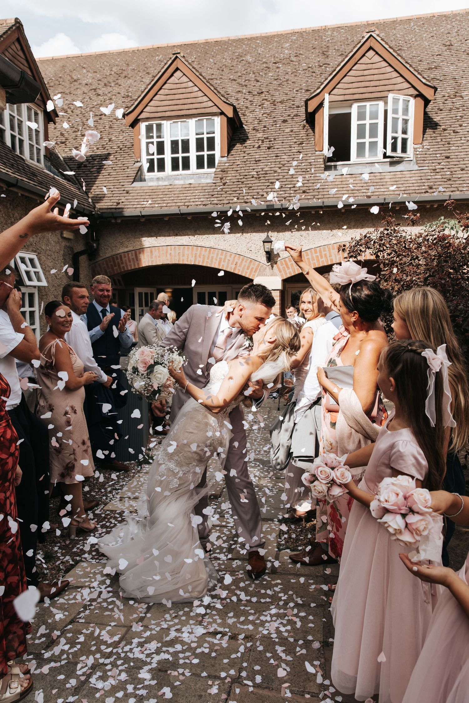 Bride and groom share a kiss surrounded by guests throwing white confetti outside a rustic brick wedding venue.