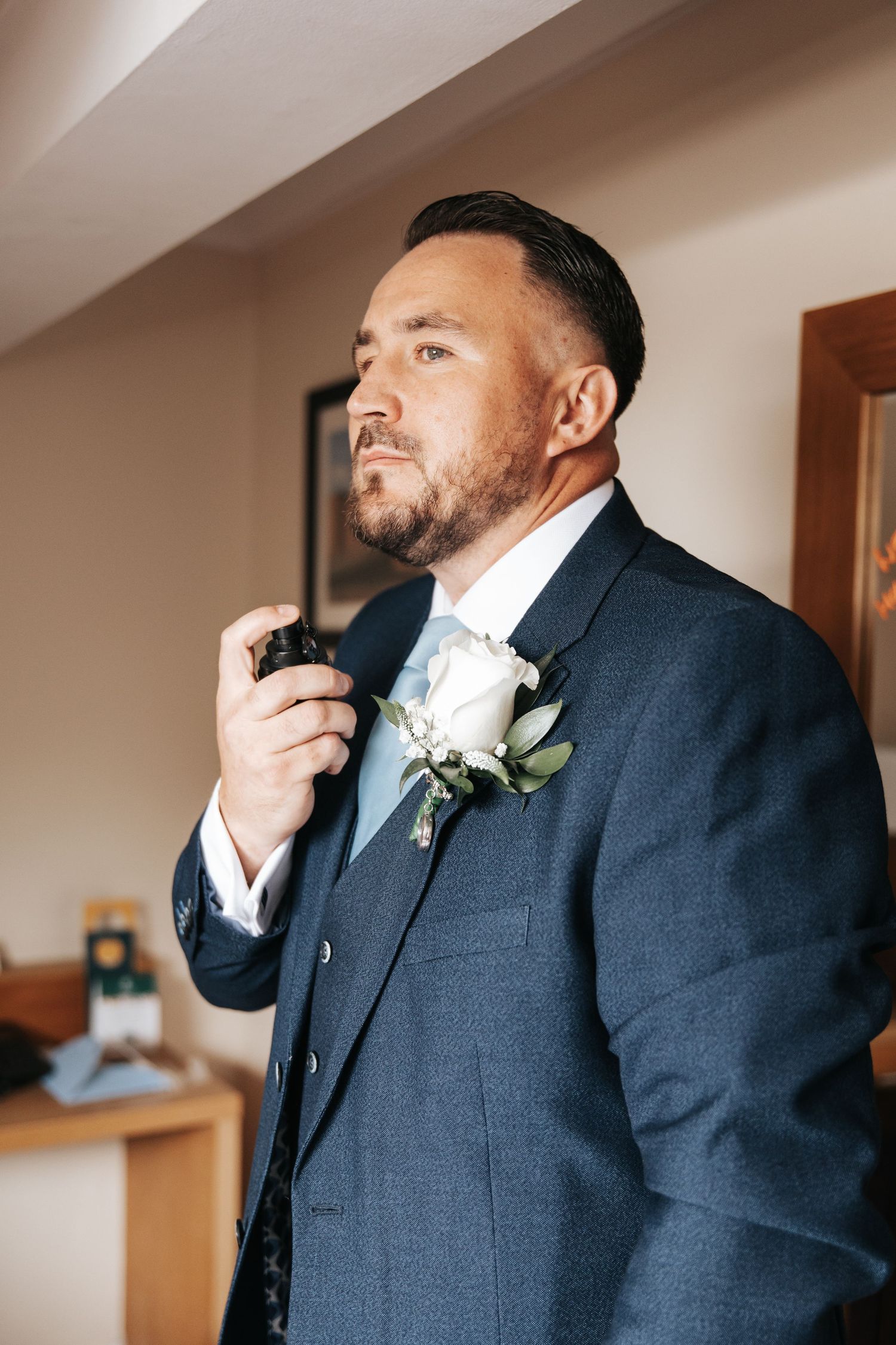 A well-dressed groom in a navy blue suit adjusts his tie, wearing a white rose boutonniere, preparing for his wedding day.