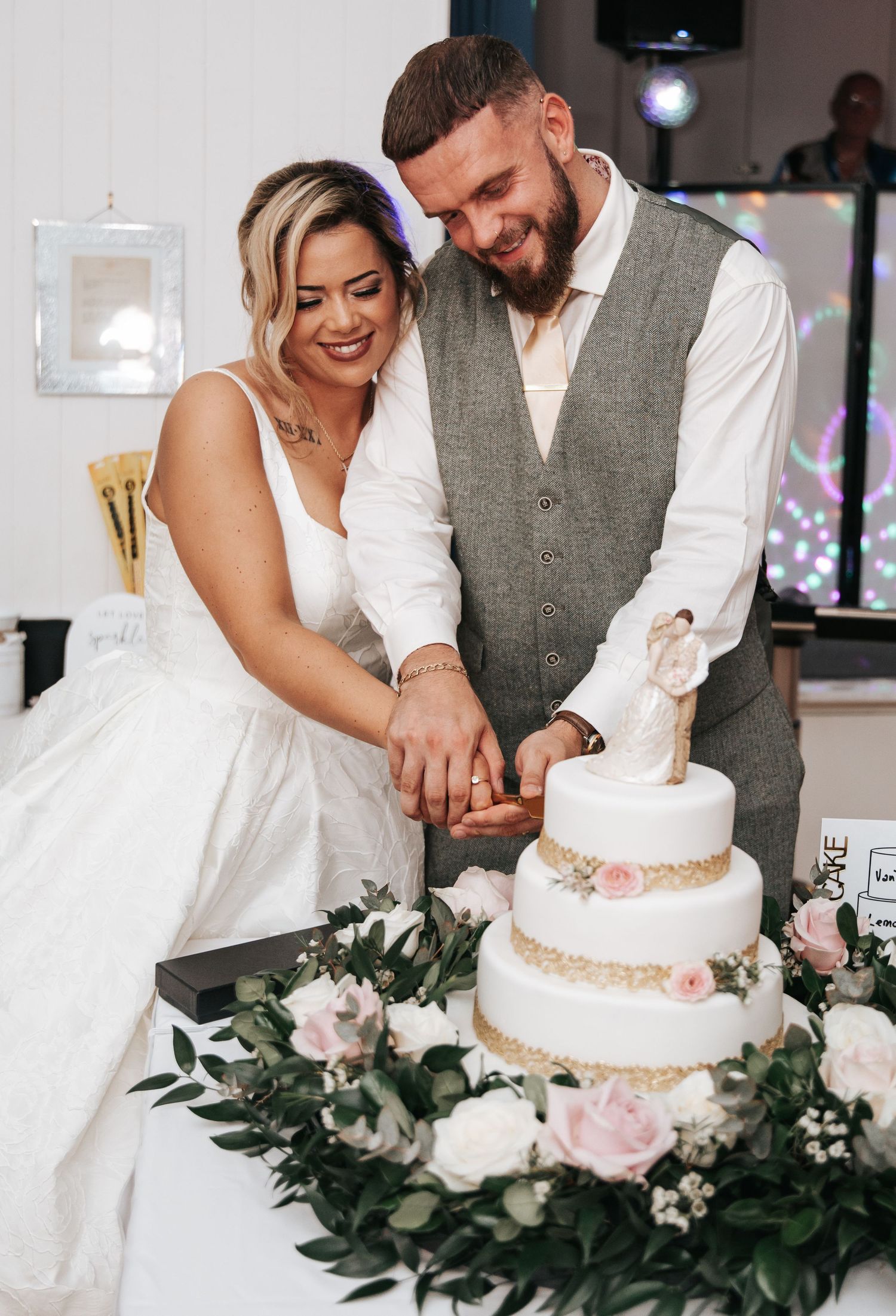 Bride and groom cutting their three-tier white wedding cake decorated with pink roses and greenery at their reception.