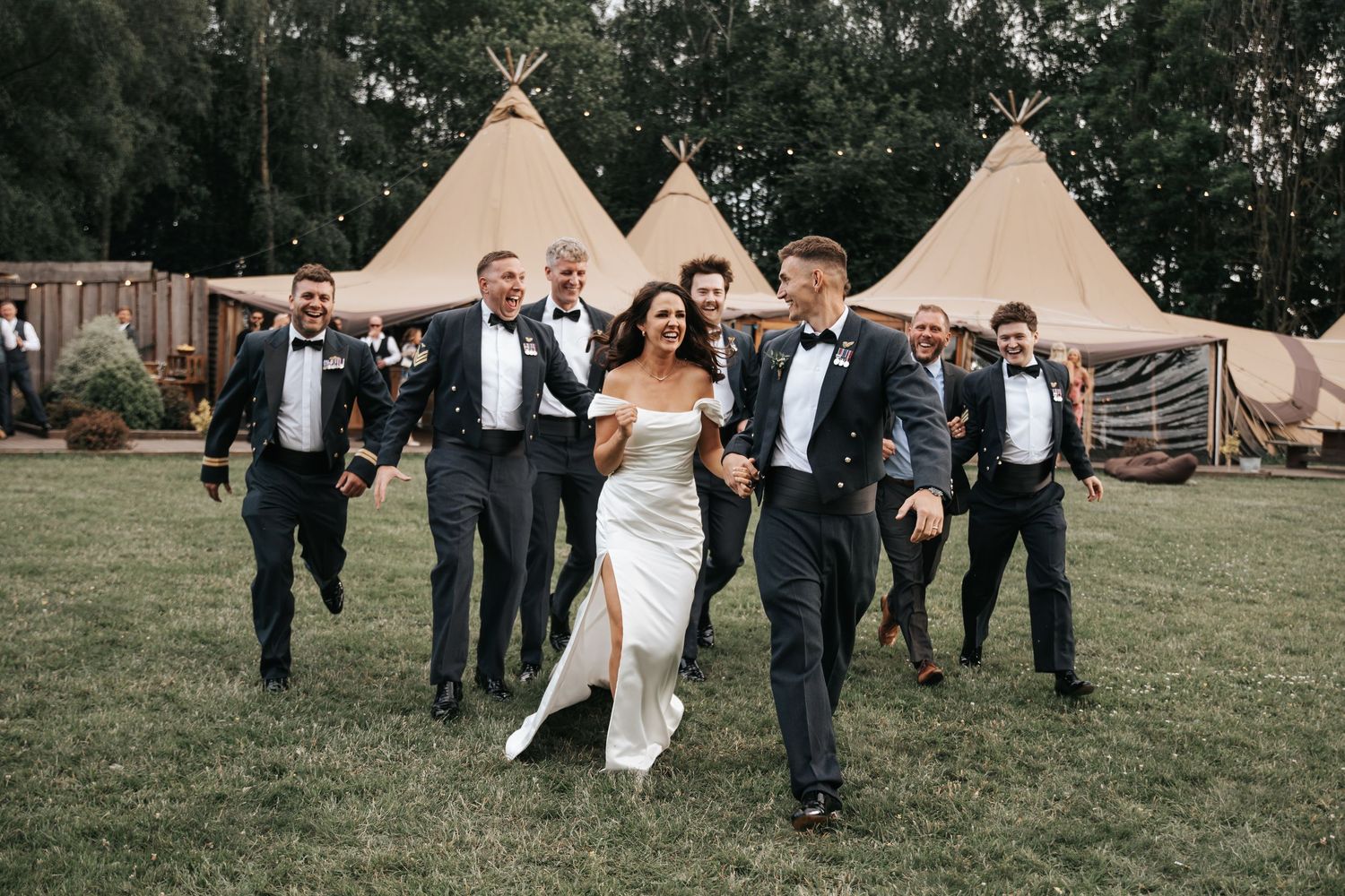 Bride and groom laugh with groomsmen in tuxedos outside tipi wedding venue on lush green lawn.