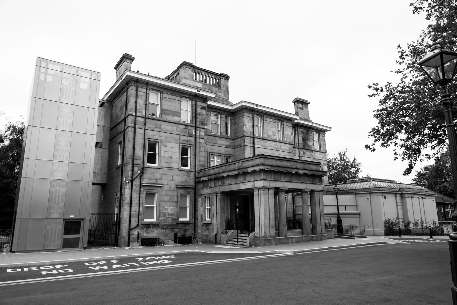 Black and white photo of a historic stone mansion with modern glass extension, featuring classical architecture and manicured grounds.
