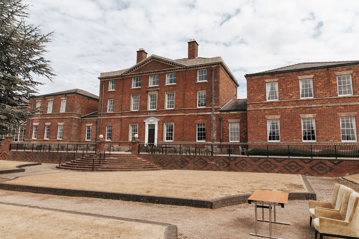 Grand Georgian red brick manor house with symmetrical facade, white sash windows, and formal grounds under cloudy skies.