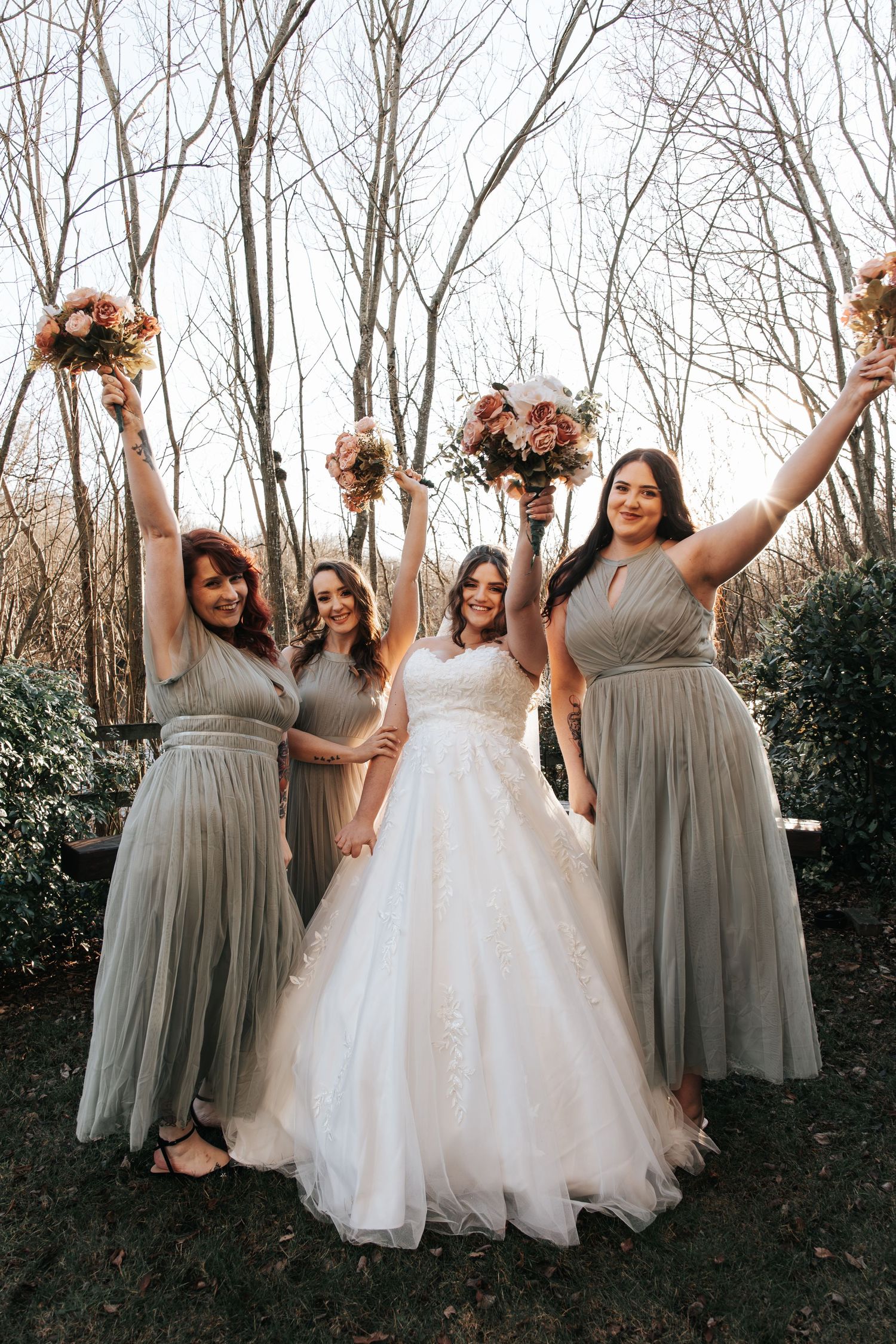 Bridal party in grey dresses celebrates with raised bouquets in winter woodland setting.