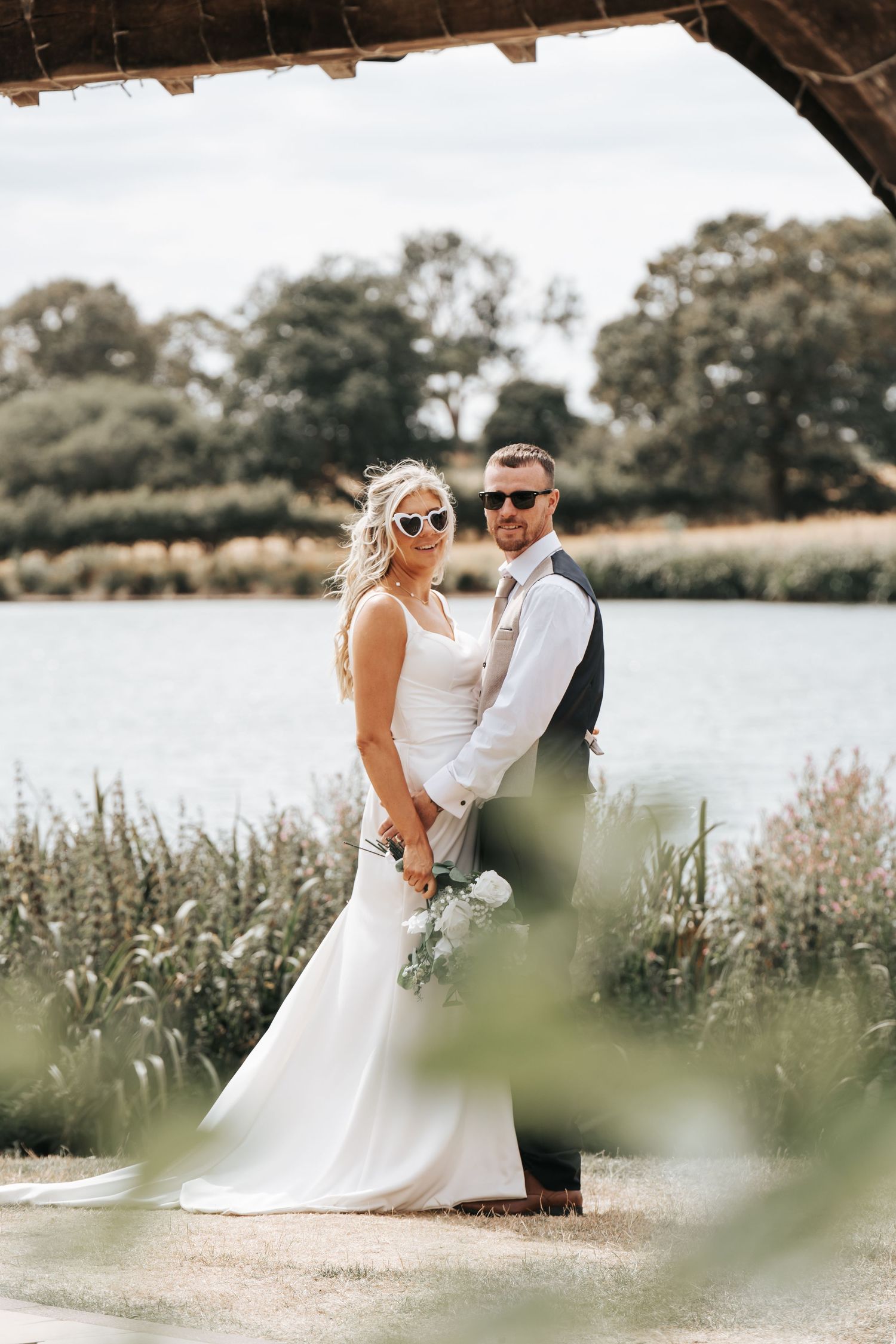 Bride in white gown and groom in vest pose by a lake, both wearing sunglasses, holding a bouquet outdoors.