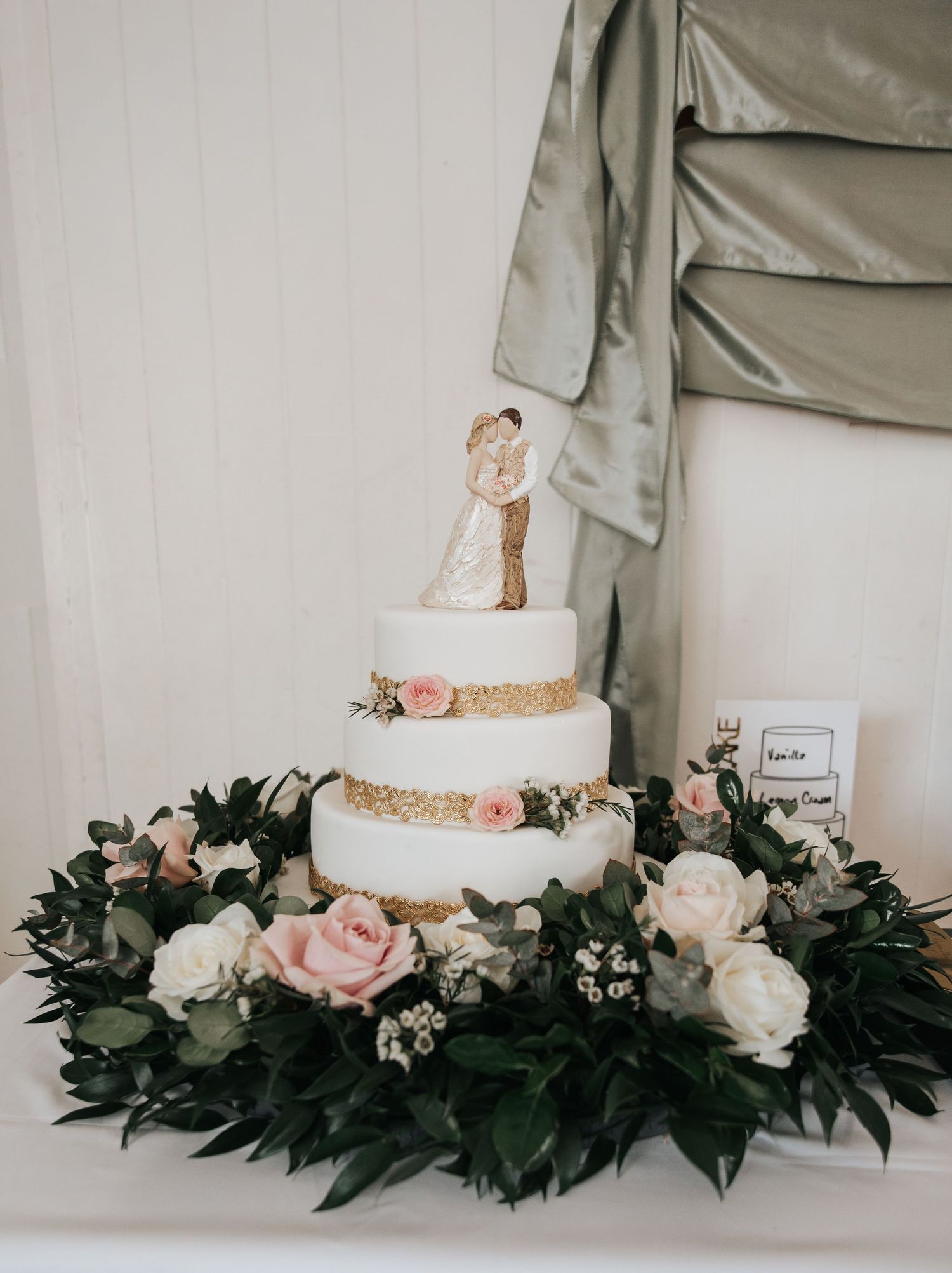 Three-tier white wedding cake with gold accents, pink roses, and a bride and groom topper surrounded by a lush floral wreath.
