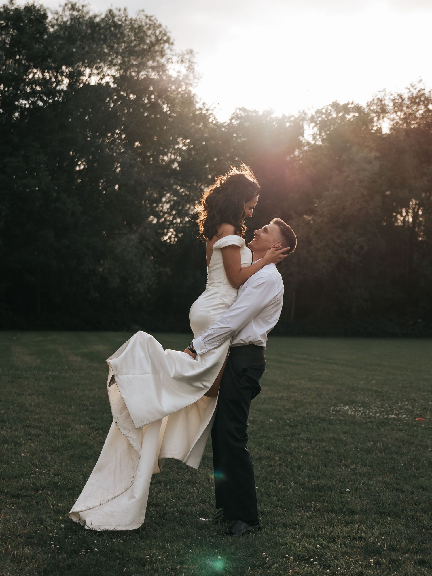 Groom lifting bride in white wedding dress at golden hour in lush green field with sunlight streaming through trees.