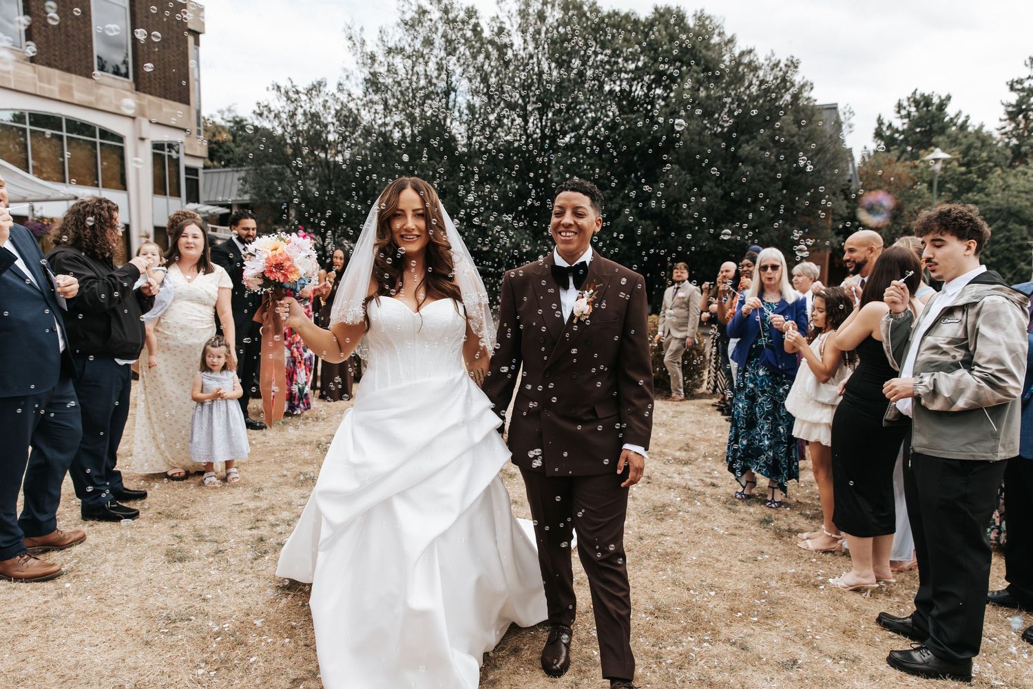 Joyful newlywed couple walking through confetti shower surrounded by cheering wedding guests outdoors.