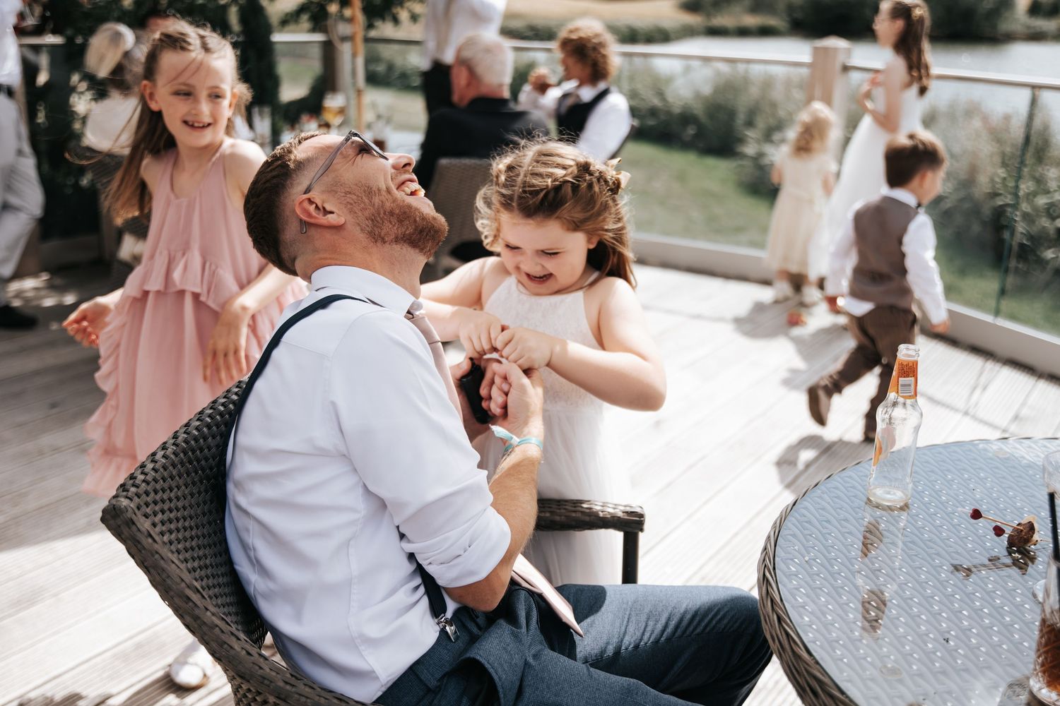 A laughing groom sits outdoors as two young girls in formal dresses playfully adjust his tie at a wedding reception.