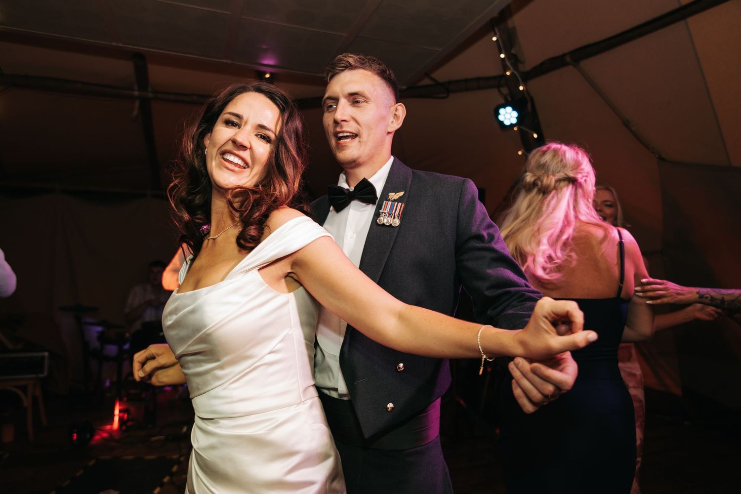 Bride and groom dancing joyfully at their wedding reception in a white gown and military dress uniform.