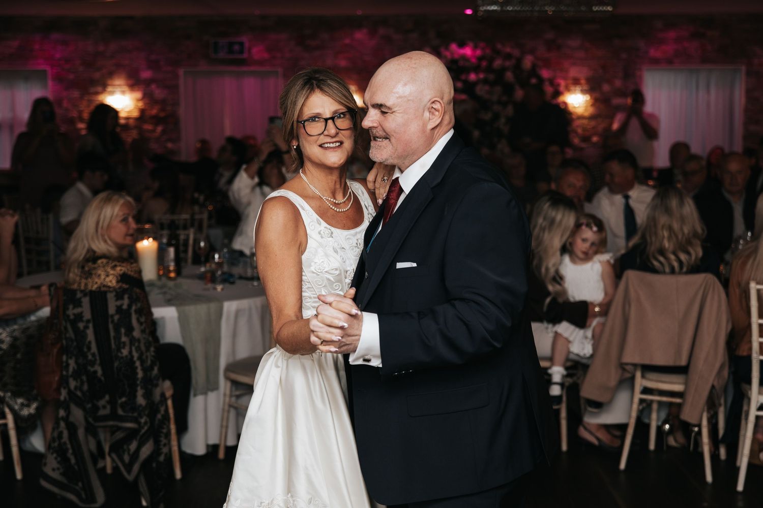 Couple sharing a dance at their wedding reception in an elegantly lit venue with guests seated in background.