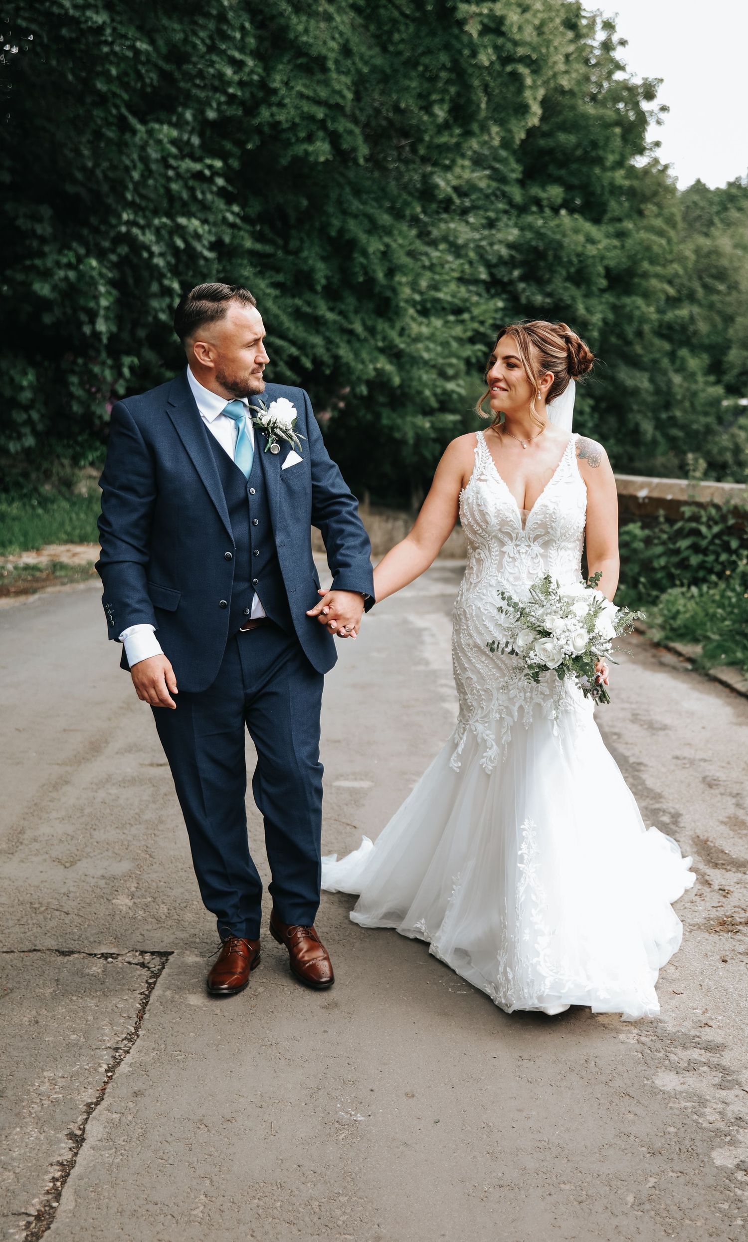 A bride and groom walk hand in hand outdoors, she in a lace mermaid gown, he in a navy blue three-piece suit.