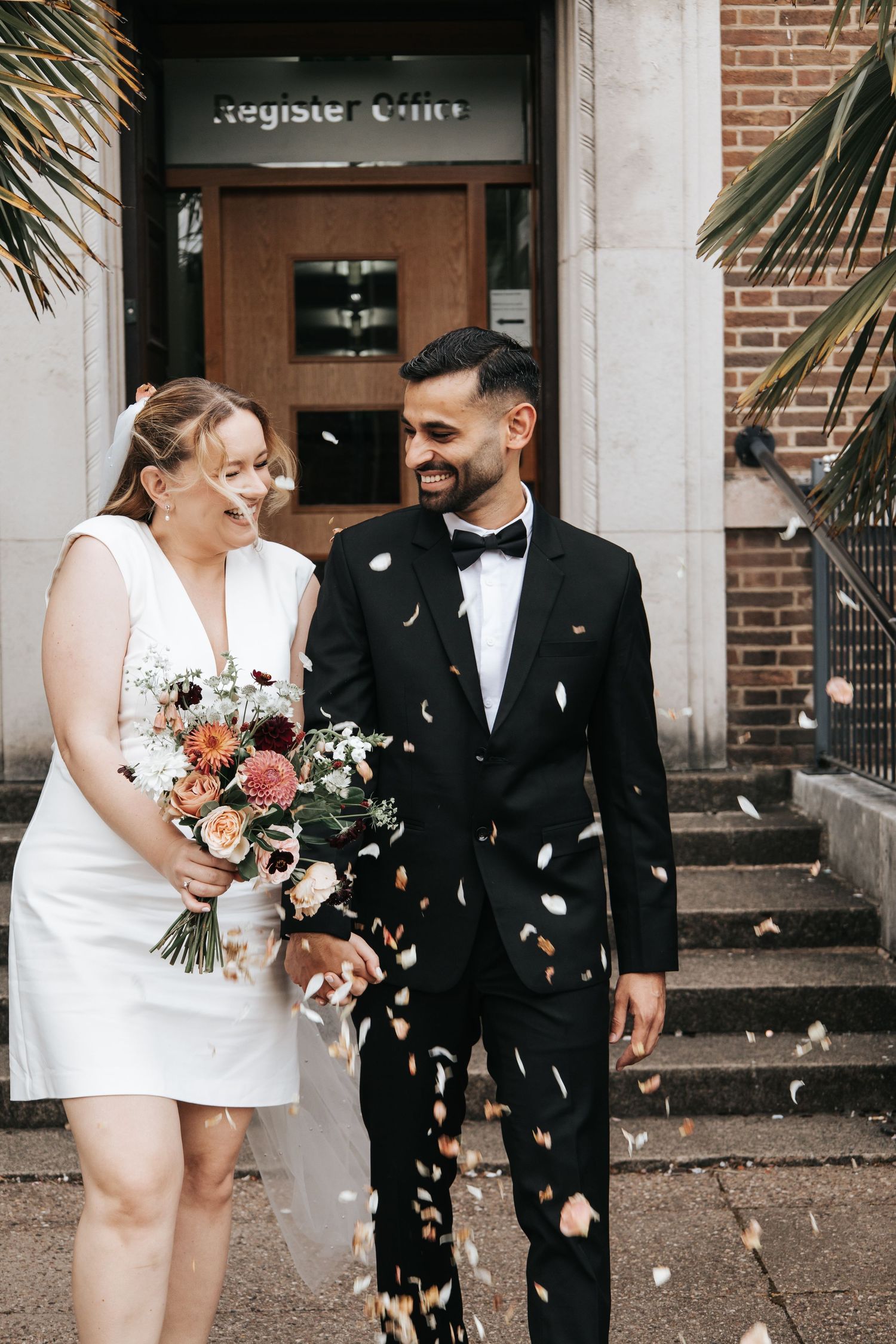 Newlyweds laughing and celebrating with flower petals outside a brick building.