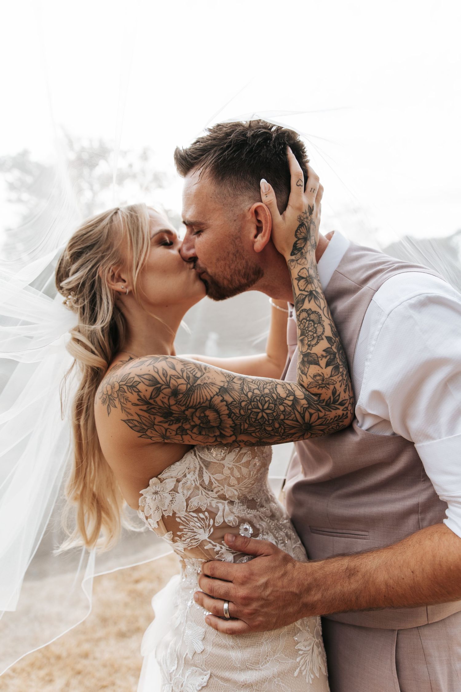 A bride and groom share a romantic kiss on their wedding day, with the bride's veil flowing and the groom's tattooed arms embracing her.
