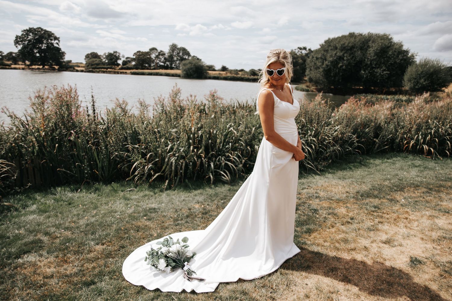 Bride in elegant white wedding gown with train stands by a serene lakeside, holding a bouquet on a sunny day.