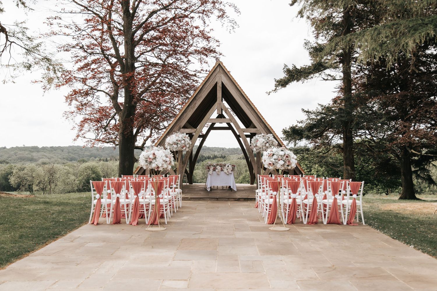 Elegant outdoor wedding ceremony setup with white chairs and floral arrangements under a wooden arch pavilion surrounded by trees.