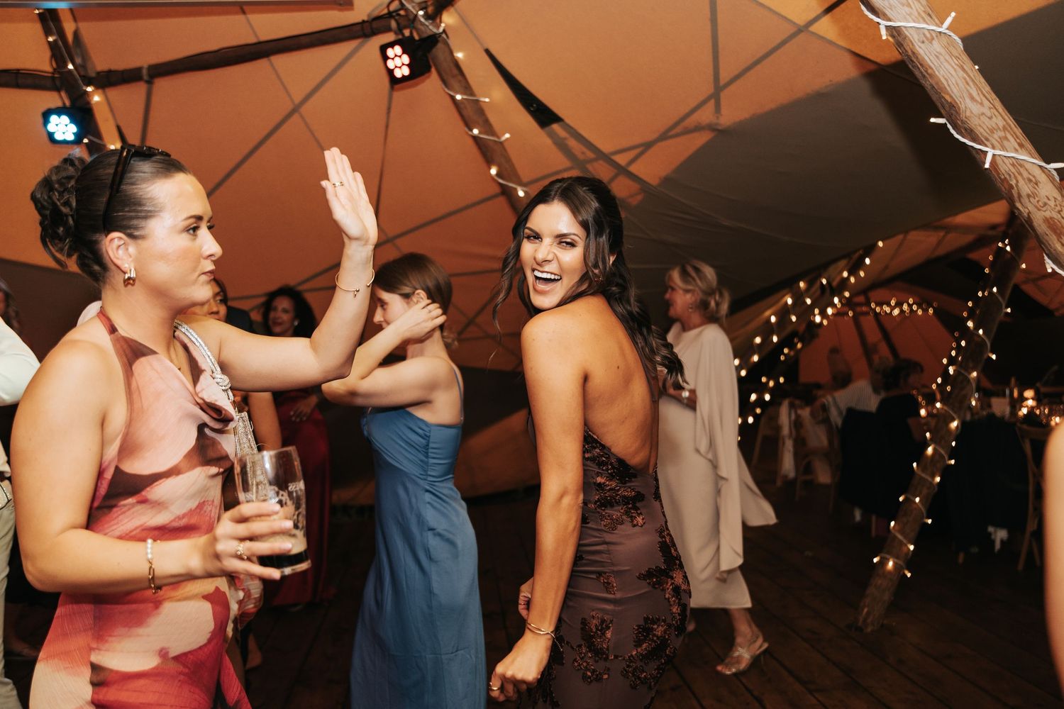 Guests dancing joyfully at an elegant wedding reception under a tent with warm string lights.