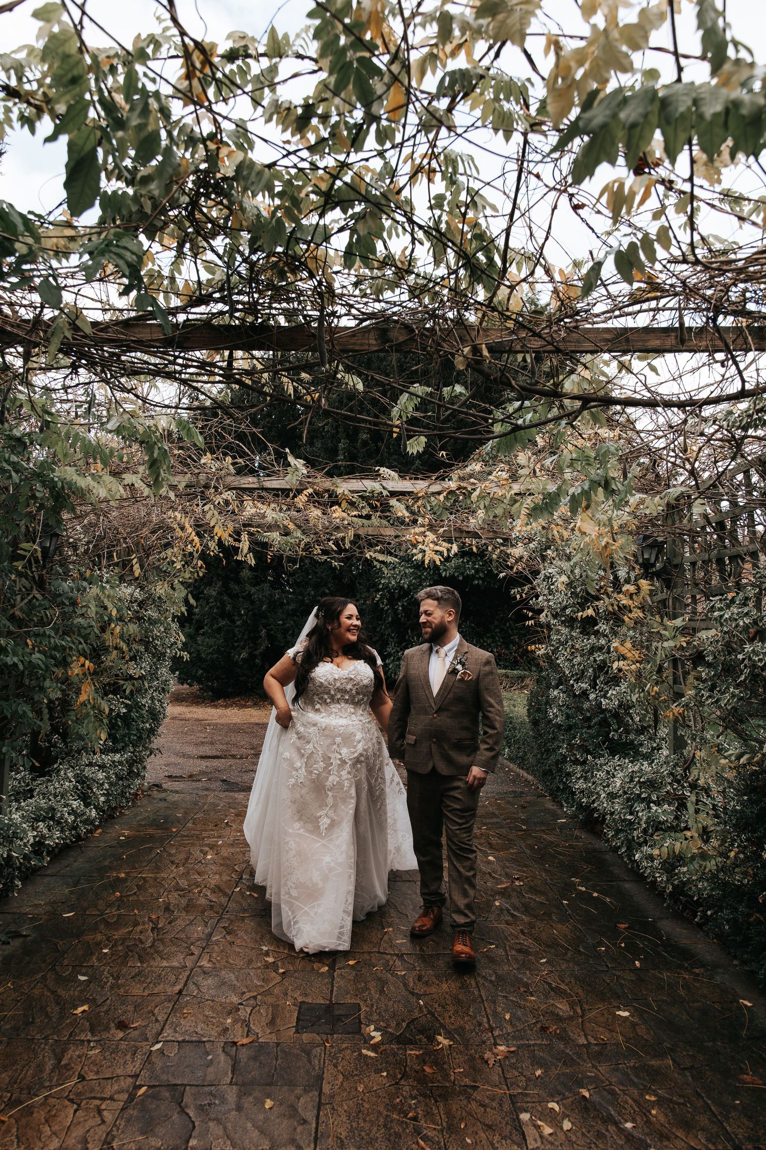 Wedding couple walking through garden archway covered in autumn foliage.