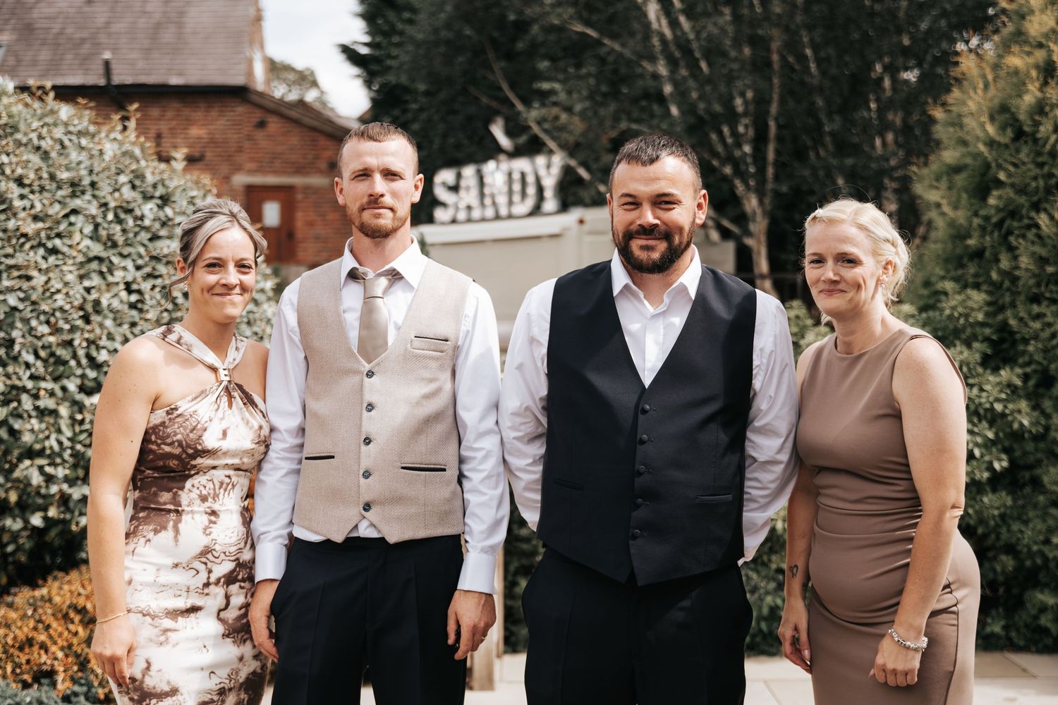 Two men in formal attire and two women in elegant dresses pose together outdoors at a wedding venue.
