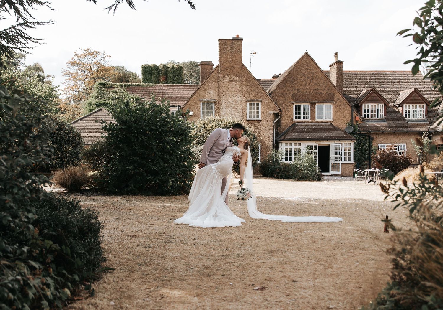 Bride and groom share a romantic dip kiss in garden of charming English countryside manor house wedding venue.