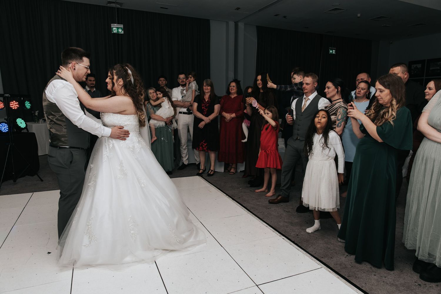 A couple shares their first dance at a wedding reception surrounded by guests on a white dance floor.