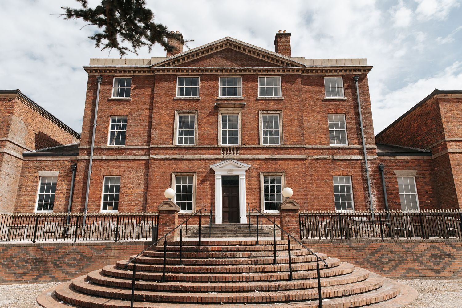 A grand Georgian red brick manor house with symmetrical windows, decorative pediment, and curved stone steps leading to the entrance.