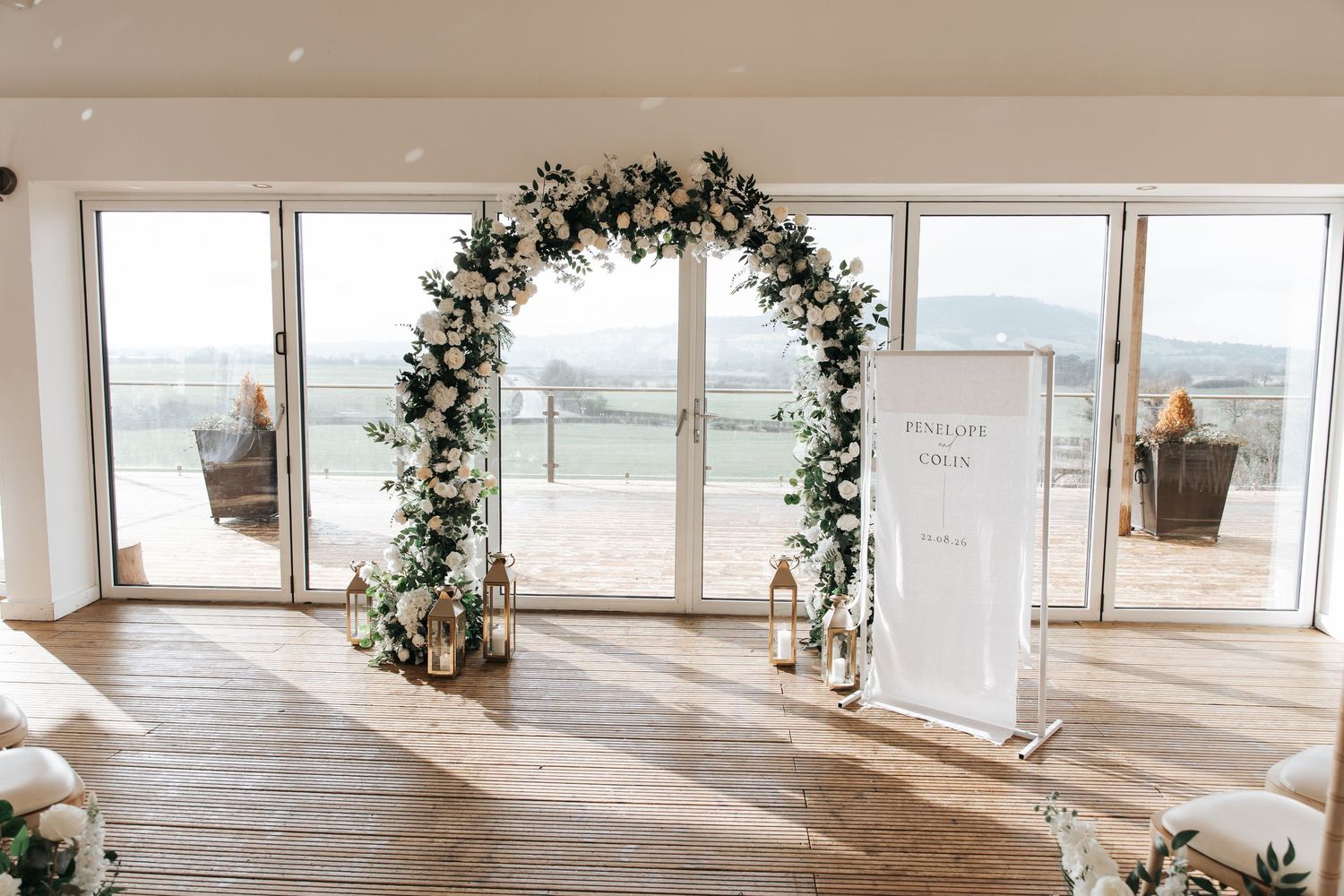 Elegant wedding ceremony arch adorned with white flowers and greenery, with candles and a welcome sign on a wooden floor.