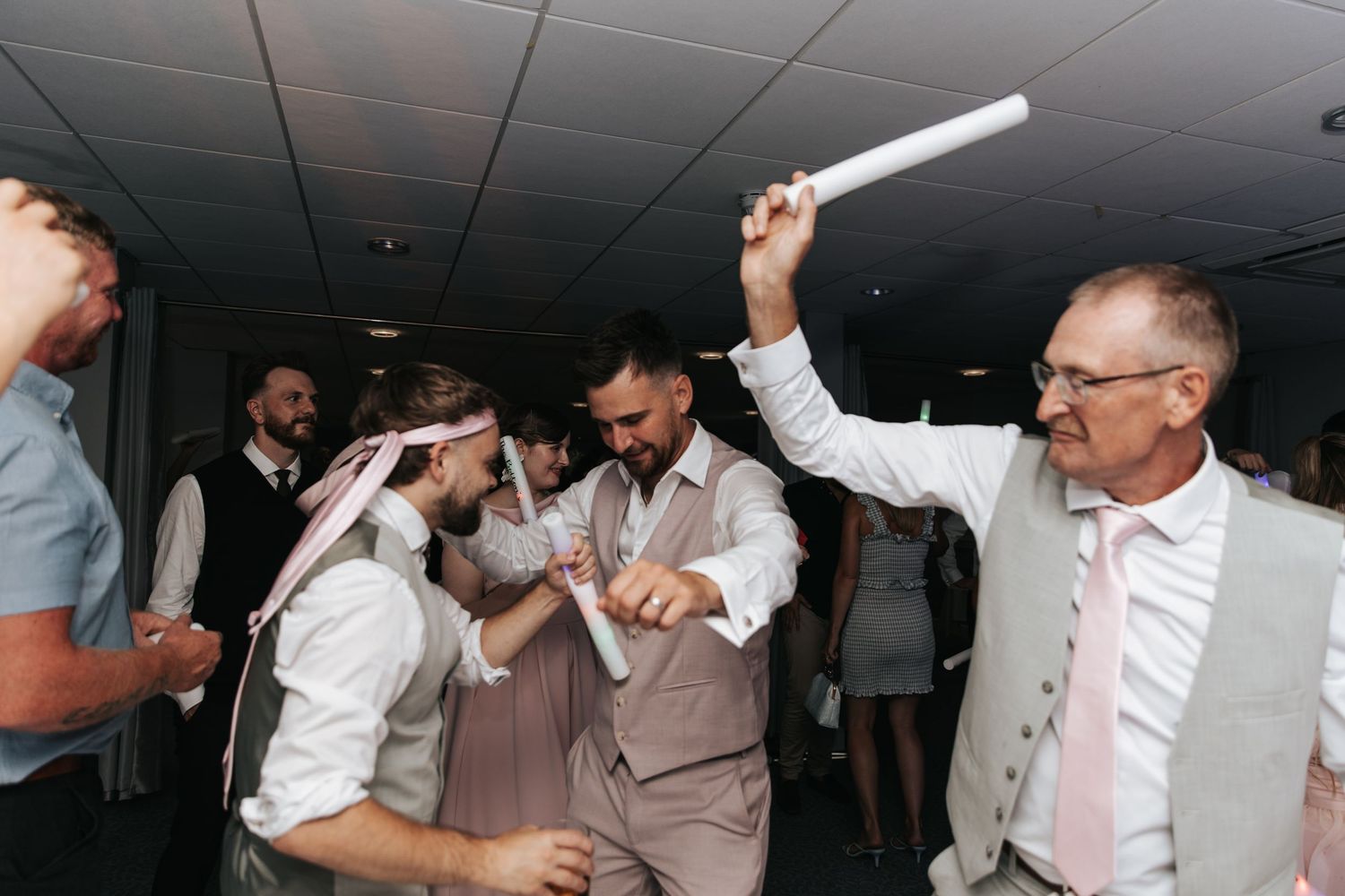 Men dancing energetically at a wedding reception, one holding a rolled paper aloft in a festive indoor celebration.