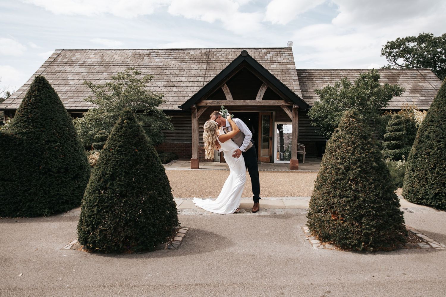 Bride and groom share a romantic kiss surrounded by topiary trees outside a rustic barn wedding venue.