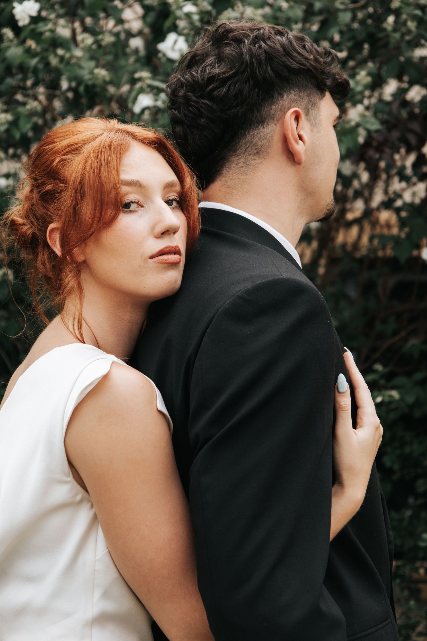 Romantic wedding portrait showing couple in white dress and black suit embracing against floral backdrop.